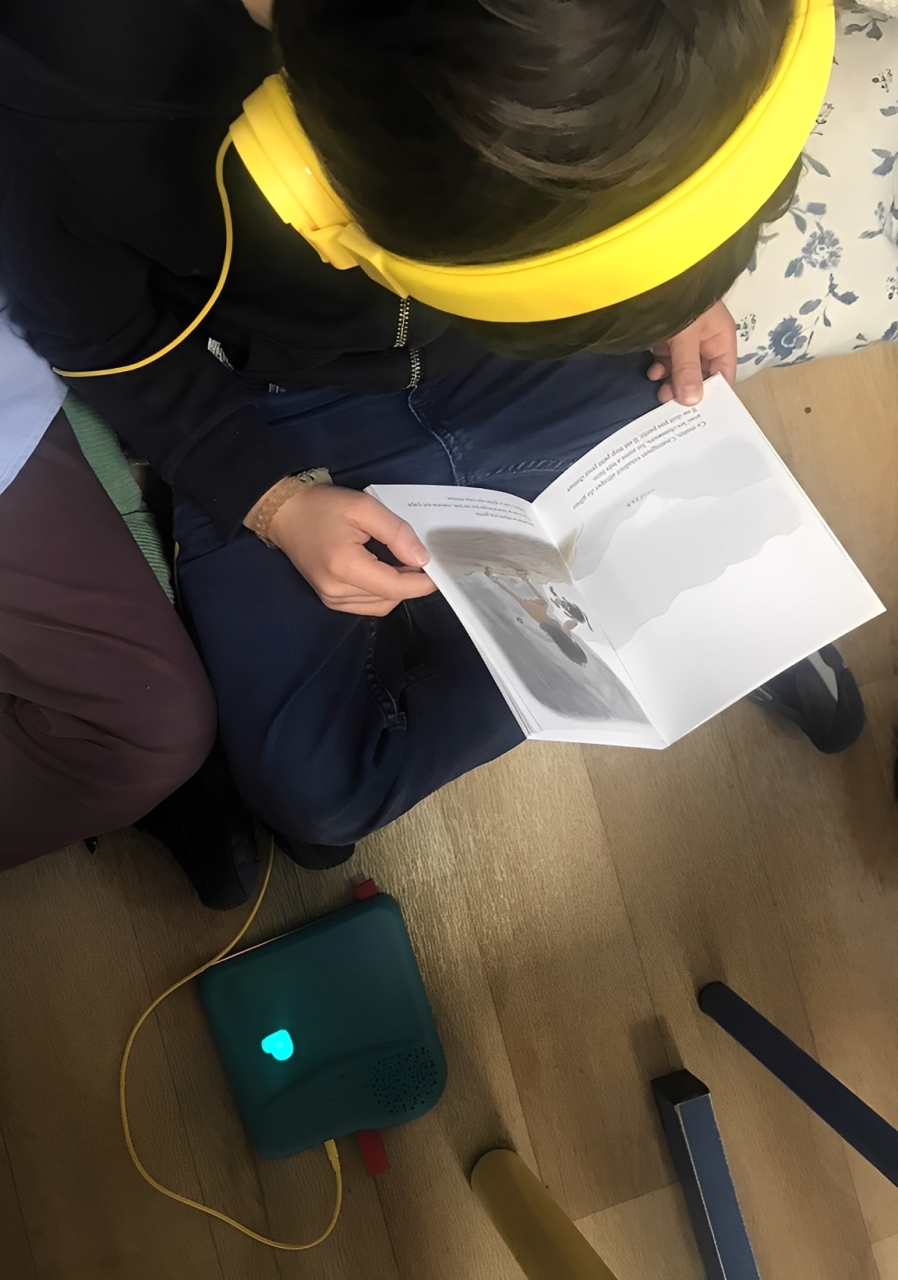 Élève assis au sol lisant un album jeunesse avec un casque audio, relié à un Bookinou.
